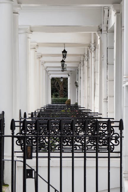 A narrow outdoor corridor with white-painted wooden columns and beams forming a covered walkway. The floor is painted black and appears clean, with black wrought iron railings lining the sides. Hanging lantern-style light fixtures are attached to the ceiling, and the corridor opens to greenery at the far end, including trees and shrubs, indicating a garden or residential outdoor space. The scene is well-lit with natural daylight, emphasizing the pristine condition of the surfaces as part of professional cleaning and maintenance services by Kensington Carpet Cleaning, aligned with surface cleaning and sanitisation themes for end of tenancy and domestic cleaning purposes.