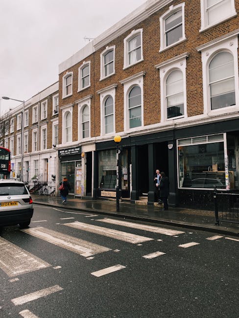 Exterior view of a multi-story commercial and residential building on High Street Kensington, with large glass display windows on the ground floor, some of which display signage for retail spaces. The building features white window frames, brick walls, and decorative architectural details, with a black awning over the storefront. The street is wet, indicating recent rain, and there are pedestrians crossing at a zebra crossing and walking along the sidewalk, some holding umbrellas. A white van and a red bus are visible on the street. The scene is illuminated by overcast sky and street lighting, creating a muted but clear urban setting relevant to domestic and commercial surface cleaning and maintenance awareness for Kensington Carpet Cleaning.