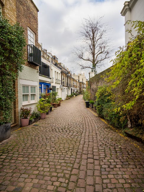 A narrow cobblestone alleyway in W8 postcode, lined with white and pastel-colored residential buildings featuring bay windows and small balconies. Potted plants and greenery are placed along the building bases, creating a tidy and well-maintained appearance. A leafless tree stands at the end of the alley, under an overcast sky, with diffused natural light illuminating the scene. The alley appears clean and free of debris, exemplifying a quiet, residential area in Kensington. [COMPANY_NAME] specializes in surface cleaning and household sanitisation, ensuring spaces like this remain hygienic and well-presented.