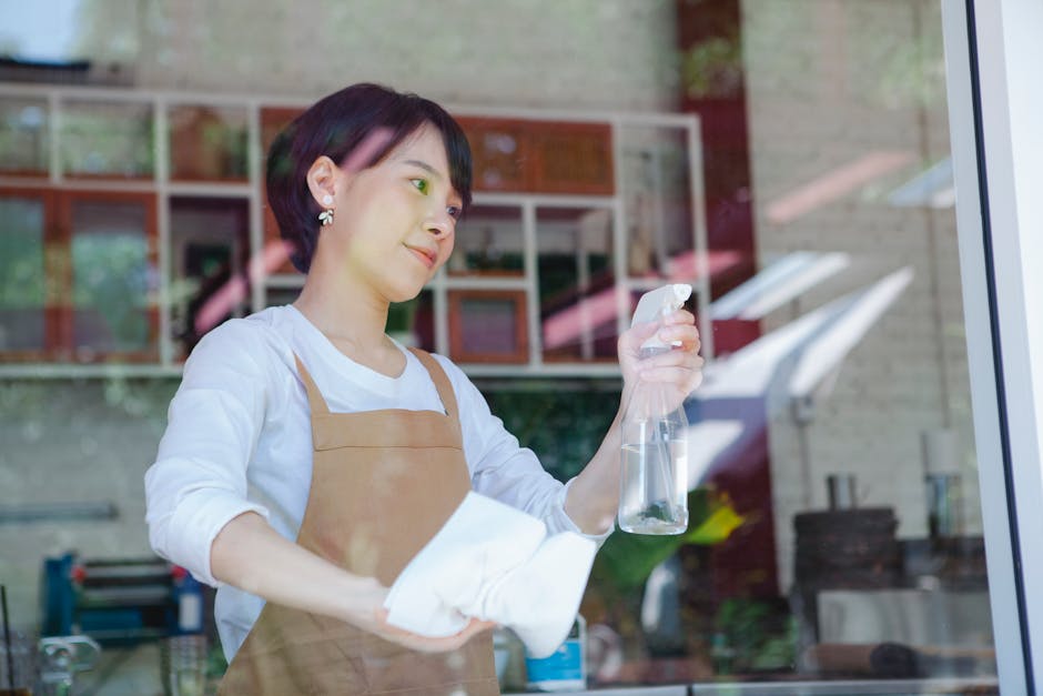 A woman dressed in a white shirt and beige apron is standing inside a commercial kitchen or restaurant, seen through a large glass window. She is holding a spray bottle filled with cleaning solution in one hand and a white cloth in the other, preparing to wipe down a surface. The background features open shelving with various kitchen items, including containers and utensils, along with stainless steel appliances and tiled walls. Natural light illuminates the space, highlighting the clean and well-maintained surfaces, with a focus on hygiene and sanitation as part of surface cleaning and general maintenance, consistent with Kensington Carpet Cleaning's cleaning services for Kensington restaurants and small businesses.