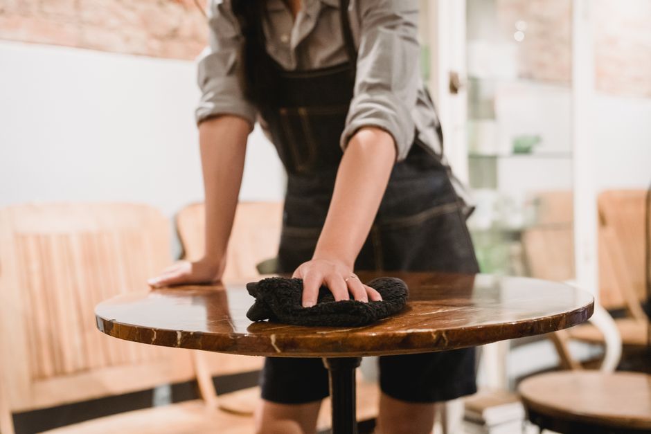 A woman with short, curly red hair wearing a white shirt and beige apron is bent over, cleaning the floor of a restaurant or café with a mop. She is positioned near a wooden table with four metal legs, which is upside down, indicating floor cleaning activity. The background features a dark green wall with a display case filled with pastries and baked goods, along with a glass cake stand, cups, and kitchen items on shelves, under soft overhead lighting. The scene emphasizes surface cleaning and hygiene practices, consistent with deep cleaning in a commercial restaurant or small business setting, as part of Kensington Carpet Cleaning's services.