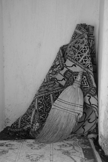 A broom with a long handle and straw bristles leaning against a pile of folded patterned rugs resting on a light-colored tiled floor, next to a plain white wall in a corner of a room. The rugs feature geometric and tribal designs, with some partially unfolded. The area appears clean, with natural lighting illuminating the scene, indicative of domestic surface cleaning and maintenance efforts. The image is associated with Kensington Carpet Cleaning, emphasizing professional rug cleaning tips for maintaining hygiene and appearance. The scene captures an organized space prepared for deep cleaning or routine household maintenance.