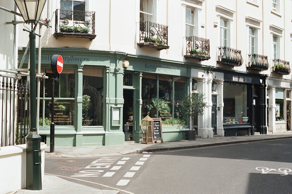 Exterior view of a multi-story building housing Kensington Flowers and an art gallery on Gloucester Road. The ground floor features large glass windows showcasing plants and artwork. The building's facade is painted in pastel green and white, with black wrought iron balconies on the upper floors. The sidewalk includes a black bollard, a chalkboard sign advertising flowers, and a small potted tree near the entrance. The street has a designated bike lane, and a no-entry traffic sign is visible on the left side under bright, natural daylight, with no visible dirt or litter on the surfaces or pavement.