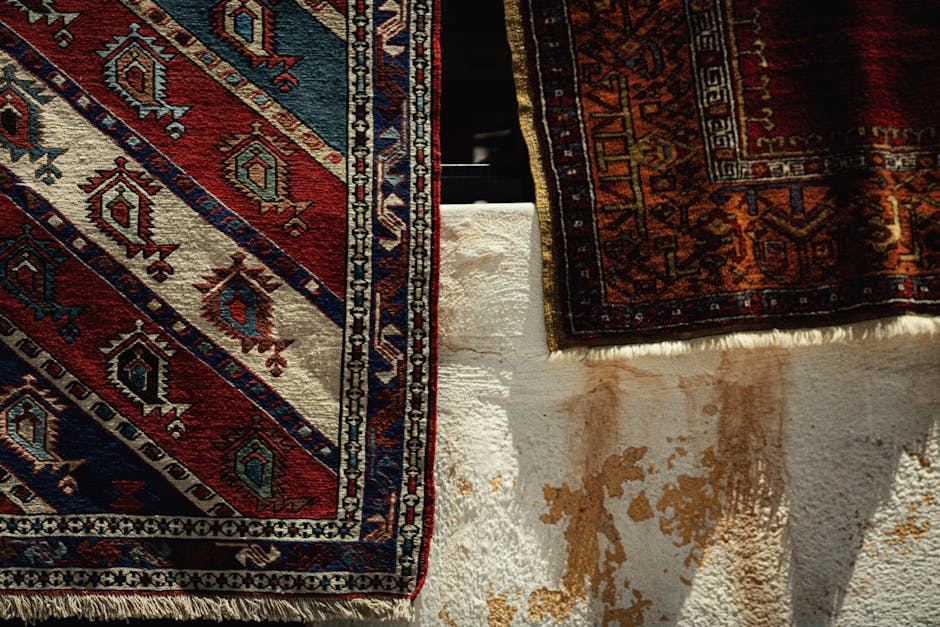 A broom with a long handle and straw bristles leaning against a pile of folded patterned rugs resting on a light-colored tiled floor, next to a plain white wall in a corner of a room. The rugs feature geometric and tribal designs, with some partially unfolded. The area appears clean, with natural lighting illuminating the scene, indicative of domestic surface cleaning and maintenance efforts. The image is associated with Kensington Carpet Cleaning, emphasizing professional rug cleaning tips for maintaining hygiene and appearance. The scene captures an organized space prepared for deep cleaning or routine household maintenance.