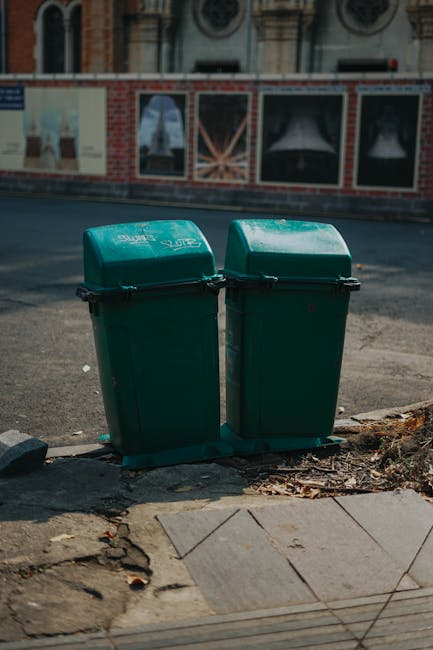 Two green outdoor waste bins with black lids are positioned side by side on a concrete sidewalk next to a cracked and chipped pavement area. The bins are marked with the logo of Kensington and Chelsea council for waste disposal. In the background, a brick building with arched windows and decorative stone details is visible, illuminated by daylight. The scene is clean, with no visible trash around the bins, indicating good maintenance. The image emphasizes the importance of proper waste disposal, relevant to the guidelines outlined on the Kensington and Chelsea council rules for waste disposal cleaning, as part of professional cleaning services offered by Kensington Carpet Cleaning for maintaining hygienic outdoor environments.