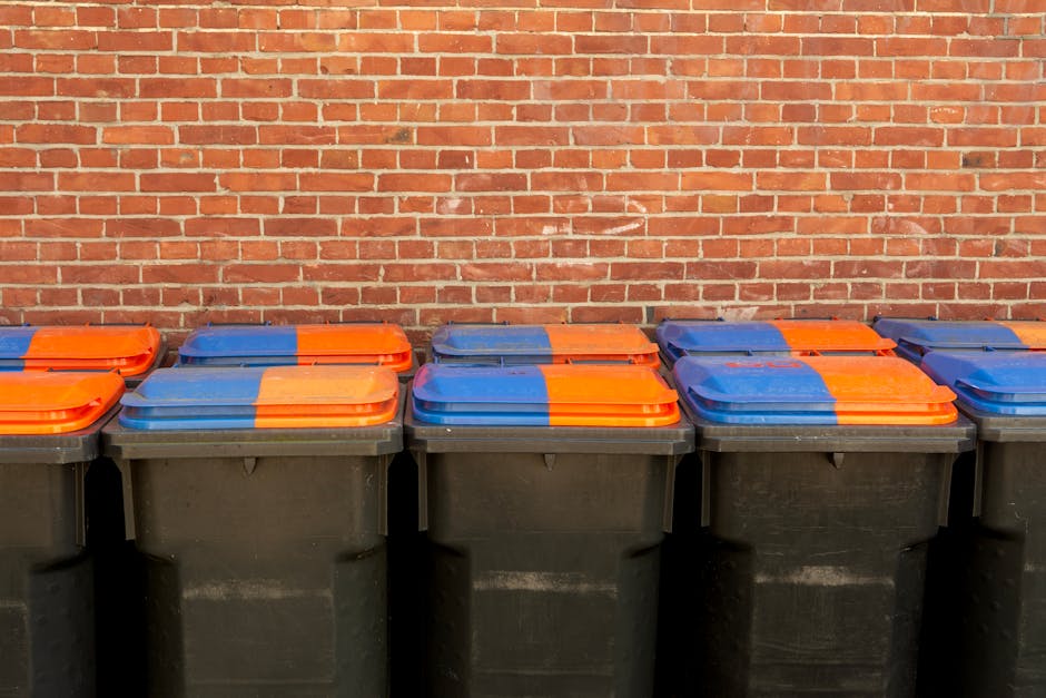 A row of black waste bins with closed lids, featuring orange and blue color-coded covers, positioned outdoors against a red brick wall. The bins are uniformly aligned, with the lids stacked flat on top, and are part of waste disposal management following Kensington and Chelsea council rules. The lighting is natural, highlighting the clean, well-maintained condition of the bins and the brick surface. This setting reflects proper waste segregation and sanitation practices, as promoted by Kensington Carpet Cleaning for hygiene and environmental responsibility.