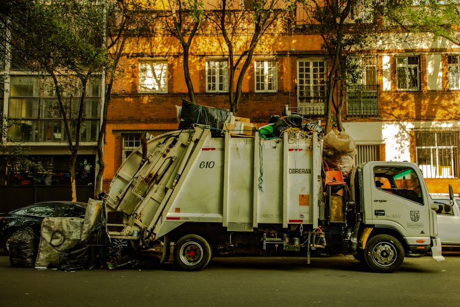 Two green outdoor waste bins with black lids are positioned side by side on a concrete sidewalk next to a cracked and chipped pavement area. The bins are marked with the logo of Kensington and Chelsea council for waste disposal. In the background, a brick building with arched windows and decorative stone details is visible, illuminated by daylight. The scene is clean, with no visible trash around the bins, indicating good maintenance. The image emphasizes the importance of proper waste disposal, relevant to the guidelines outlined on the Kensington and Chelsea council rules for waste disposal cleaning, as part of professional cleaning services offered by Kensington Carpet Cleaning for maintaining hygienic outdoor environments.