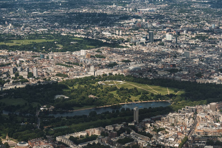 An aerial view of Kensington Gardens in London, showing the lush green lawns and the reflective waters of the Serpentine River, surrounded by urban buildings and city streets. The scene highlights the contrast between the natural parkland and the dense cityscape beyond. The park's pathways and scattered trees are visible amid the well-maintained grass, with the city skyline featuring various modern and historic structures. Natural light illuminates the scene, emphasizing the cleanliness and maintained appearance of the area, consistent with the expertise of Kensington Carpet Cleaning in surface cleaning and maintenance of both outdoor and indoor environments.