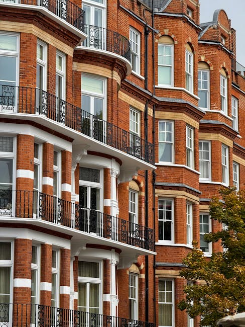 An exterior view of a multi-story residential building with red brick walls, white decorative bands, and black metal balconies. The building features large, evenly spaced double-hung sash windows with white frames, and a small tree with green and orange leaves is visible in the foreground, suggesting autumn. The sunlight illuminates the facade, highlighting the cleanliness and well-maintained appearance of the surfaces. The image exemplifies high-quality exterior surface cleaning and maintenance, consistent with the services offered by Kensington Carpet Cleaning, as part of their domestic cleaning and surface sanitation expertise.
