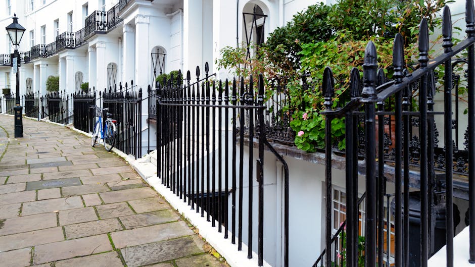 A residential street in Kensington featuring a paved sidewalk with rectangular stone slabs alongside white townhouse buildings with black wrought iron railings and balconies. A blue bicycle is parked upright against the railing near the sidewalk. The exterior walls of the buildings are clean, with potted plants and greenery visible on the balconies and near the windows. Bright natural light illuminates the scene, highlighting the well-maintained appearance of the outdoor areas, consistent with professional cleaning standards. This image exemplifies a tidy, well-kept residential environment, aligning with the service quality offered by Kensington Carpet Cleaning. No visible clutter or dirt suggests ongoing or recent surface cleaning or maintenance.