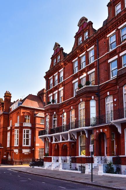 Photograph of a red brick residential building with multiple large windows on each floor, some with white frames and decorative lintels. The building features small balconies with black wrought iron railings and white supports, and a white staircase leading to the entrance on a city street. The sidewalk appears clean and well-maintained, with a clear blue sky overhead. The image is relevant to the services of Kensington Carpet Cleaning, highlighting the importance of surface cleaning and maintenance in urban residential environments, supporting their expertise in domestic cleaning and sanitisation as explained on the page titled 'Licensing and insurance for Kensington cleaners explained' at kensingtoncarpetcleaning.org.uk.