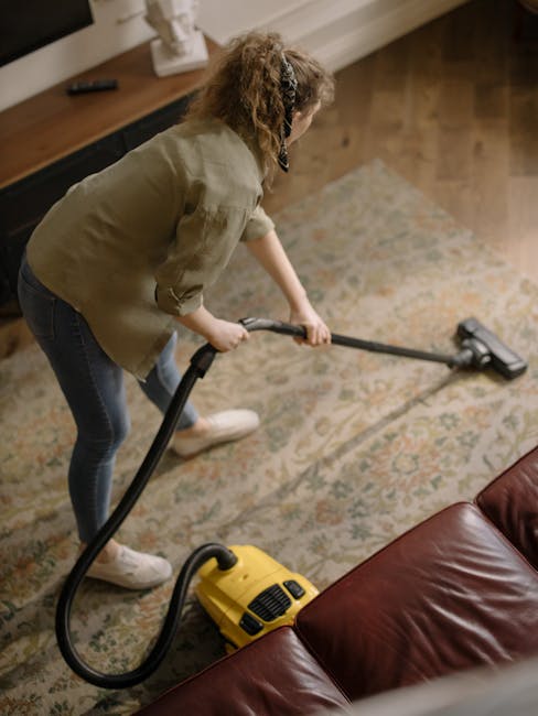 A woman dressed in casual clothing, including a beige jacket, dark jeans, and white sneakers, is using a yellow vacuum cleaner with a black hose attachment to clean a patterned area rug on a wooden floor in a living room. The rug features intricate designs and neutral tones, and the room has warm lighting. In the background, there is a wooden sideboard with a decorative item on top and a window with blinds. The scene demonstrates surface cleaning and deep cleaning processes aligned with professional carpet and upholstery sanitisation, as provided by Kensington Carpet Cleaning in Kensington W8.