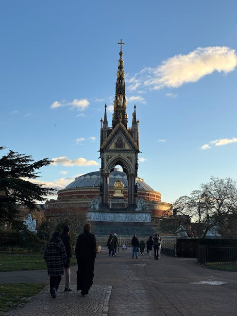 A view of the Royal Albert Hall area with a prominent ornate monument featuring intricate gothic-style detailing and a pointed spire topped with a cross, set against a bright blue sky with scattered white clouds. The monument is surrounded by a paved pathway where several visitors are walking towards it, with trees on either side and a few pedestrians visible in the foreground. The scene is illuminated by natural daylight, highlighting the details of the monument and the surrounding park setting, in an outdoor setting near the Kensington area. This image is related to the 'Royal Albert Hall area upholstery cleaning guide, KENSINGTON' and showcases an outdoor historic monument with clear weather conditions.