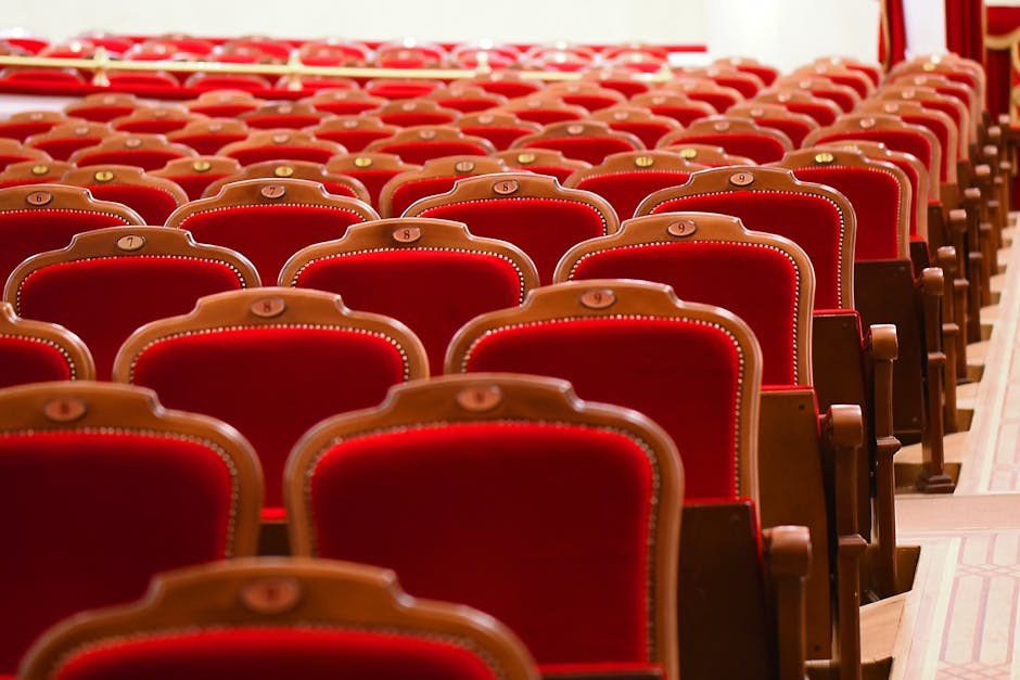 Interior view of the Royal Albert Hall showing rows of red velvet-upholstered seats with wooden armrests and brass numbering plates, arranged in a curved pattern on a wooden floor. The seats are clean and well-maintained, reflecting good surface cleaning and upkeep. The lighting is bright and evenly distributed, highlighting the rich texture of the velvet fabric and the polished wood. In the background, red curtains and part of the hall's ornate decor are visible, contributing to the elegant ambiance. The image emphasizes the importance of deep cleaning and hygienic maintenance in a prestigious venue, consistent with the services provided by Kensington Carpet Cleaning, as referenced on the page about upholstery cleaning.