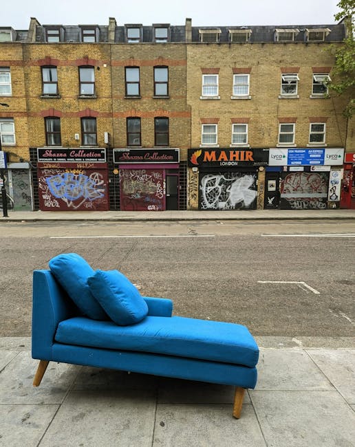 A bright blue upholstered chaise lounge with two matching cushions placed on it, positioned on a concrete sidewalk in front of a row of closed storefronts with graffiti-covered metal shutters. The background features a multi-story brick building with a mixture of red and yellow bricks, multiple windows, some with open or closed blinds, and various signage including 'Stanza Collection' and 'Mahir London'. The scene is illuminated by natural daylight, with the street appearing clean and the furniture clean and well-maintained, suggesting a focus on surface cleaning and deep cleaning services offered by Kensington Carpet Cleaning near South Kensington station.
